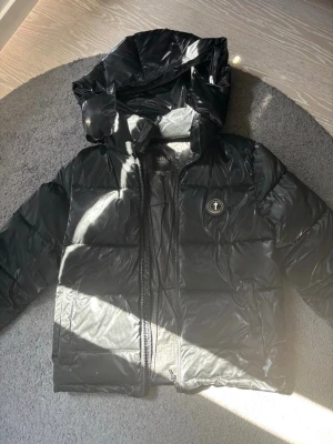 Svart pufferjacka från Trapstar - Säljer en svart pufferjacka från Trapstar med glansig finish och broderad logga på ryggen. Jackan har huva, dragkedja framtill och en rund logotyp på bröstet. Perfekt för kalla vinterdagar och riktigt streetig vibe.
