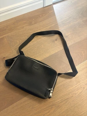 Svart axelväska från Lacoste - Snygg svart axelväska från Lacoste med präglad logotyp framtill. Väskan har justerbart axelband, silverfärgade dragkedjor och ett rymligt huvudfack med innerficka. Tillverkad i slitstarkt syntetmaterial med grön insida och diskret Lacoste-mönster, äkta självklart, köpt 2022.