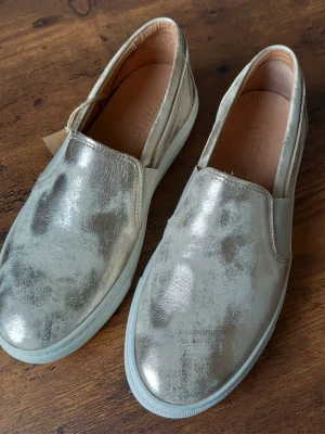 Metallic slip-on sneakers i gold från Ten Points storlek 39 - Snygga slip-on sneakers från Ten Points i goldärgat läder. Skorna har elastiska paneler på sidorna för enkel påtagning och en vit, platt sula. Perfekta för dig som vill sticka ut med en cool och modern look.