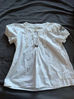 Vit blus med volanger från Odd Molly - Supersöt vit blus från Odd Molly med korta puffärmar, volangdetaljer längs knäppningen och en liten rosett framtill. Broderad fågel med hjärta på ena ärmen. Tillverkad i mjukt bomullstyg, perfekt för en chill och stilren look.