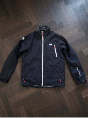  Helly Hansen jacka, perfekt skick! - Svart regnjacka från Helly Hansen med dragkedja och röda detaljer. Jackan har tre fickor med dragkedja och HH-logga på bröstet och ryggen. Materialet är vattenavvisande och perfekt för vår och höst. Jackan är aldrig använd och sitter riktigt bra i storleken M. Skriv vid minsta fråga eller fundering!