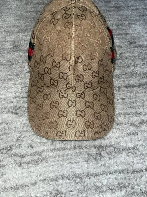Beige keps med grön och röd mönster på sidorna - Snygg beige keps med grön och röd linjer på sidorna av kepsen. Man kan ha den på vilken storlek som helst och den är bra för alla terminer. Den passar med vilken klädstil som helst.