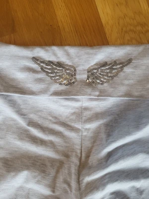 Grå mjuka loungewear-byxor med vingar - Gråa loungewear-byxor med bred midja och glittriga vingdetaljer i silver på linningen. Byxorna har raka, vida ben och är tillverkade i ett mjukt och stretchigt material som passar perfekt för chill hemma. Snygg och avslappnad stil för dig som gillar bekväma kläder.