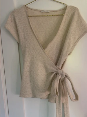 Beige omlottopp med knyt - Snygg beige omlottopp med knyt i sidan och korta ärmar. Säljer för att den inte kommer till användning.