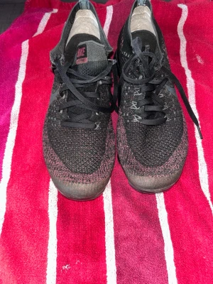 Nike svart/röd Flyknit Vapormax - Nike Vapormax sneakers i svart och rött med stickad Flyknit-överdel och genomskinlig, segmenterad sula. Skorna har svarta skosnören och diskret Nike-logga på plösen. Lätta och flexibla med modern design, perfekt för dig som gillar streetstyle. Skorna har ett par Öglor som är sönder, vid frågor eller intresse dm:)