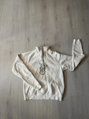 Rhinestone Hoodie - Säljer en beige hoodie med halv dragkedja och ett coolt kaninmotiv i strass på bröstet. Hoodien har långa ärmar och är gjord i mjukt sweatshirtmaterial. Perfekt för dig som gillar unika detaljer och bekväma plagg. Har tyvärr en liten fläck på ärmen, som går att se på sista bilden.