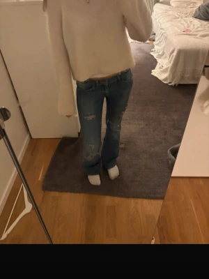 Blå bootcut jeans med slitningar - Säljer ett par blå bootcut jeans med slitna detaljer och hål på benen. Jeansen har klassisk femficksmodell, låg midja och en liten brun läderdetalj på ena bakfickan. Perfekta för en avslappnad och trendig look.