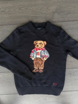 Mörkblå stickad tröja med björn från Ralph Lauren - Mörkblå stickad tröja från Ralph Lauren med ikonisk björn på bröstet. Tröjan har rund hals, ribbade muddar och RL-broderi nertill. Björnen är klädd i jeansjacka, röd scarf och bruna byxor. Perfekt för dig som gillar unika detaljer.