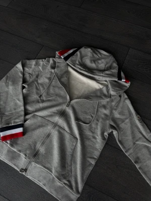 Grå hoodie med dragkedja och ränder - Snygg grå hoodie med dragkedja framtill och fickor. Huvan och ärmsluten har coola ränder i rött, vitt och blått som ger en sportig vibe. Mjuk insida och klassisk passform, perfekt till chill eller streetwear. Materialet känns som bomullsmix.