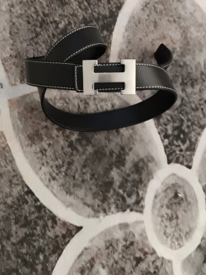 Svart skärp med silverfärgat spänne - Stilrent svart skärp med vit kontrastsöm och ett rektangulärt silverfärgat spänne. Skärpet har en klassisk design och är tillverkat i skinn, perfekt för att lyfta vilken outfit som helst.