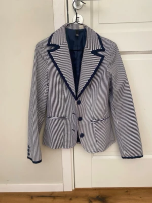 Randig kavaj med mörkblå detaljer - Snygg kavaj från H&M med smala blå och vita ränder, markerade mörkblå kanter och knappar. Klassisk krage och två fickor framtill. Perfekt för en stilren look. Fodrad insida för extra komfort.