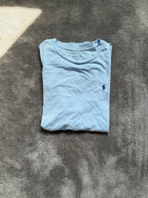 Ljusblå t-shirt från Polo Ralph Lauren - Snygg ljusblå t-shirt från Polo Ralph Lauren med klassisk rund hals och liten broderad logga på bröstet. Tillverkad i mjuk bomull och har en normal passform. Perfekt för en avslappnad och stilren look.