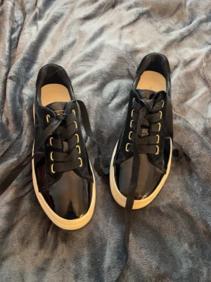 Svarta lackade sneakers från Gant - Snygga svarta sneakers från Gant i lackat material med breda svarta skosnören och guldfärgade öljetter. Skorna har vit sula och låg profil, perfekt för en trendig och stilren look.