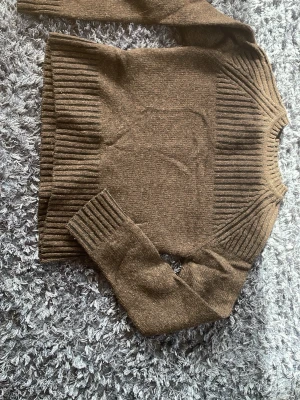 Brun stickad tröja från Calvin Klein - Snygg brun stickad tröja från Calvin Klein i storlek M. Tillverkad av 80% lammull och 20% nylon. Tröjan har breda ribbade muddar vid ärmslut, nederkant och runt halsen för en cool och modern look.