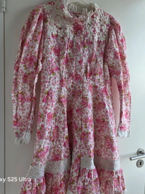 Blommig långklänning med spetsdetaljer - Romantisk långklänning med rosa och vita blommor, spetskrage och broderade blommor vid axlarna. Klänningen har långa ärmar, knappar framtill och volangdetaljer nertill. Perfekt för dig som gillar vintage vibes och unika detaljer.
