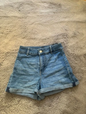 Blåa högmidjade jeansshorts - Snygga blå jeansshorts med hög midja och uppvikta benslut. Klassisk femficksmodell med och knappstängning framtill. Perfekta för varma dagar och enkel att matcha med allt.