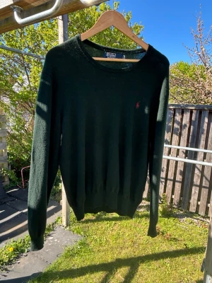 Mörkgrön tröja från Polo Ralph Lauren - Mörkgrön stickad tröja från Polo Ralph Lauren med rund halsringning och diskret röd logga broderad på bröstet. Långärmad modell med ribbade muddar vid ärmslut och nederkant. Perfekt för lager-på-lager-stil.