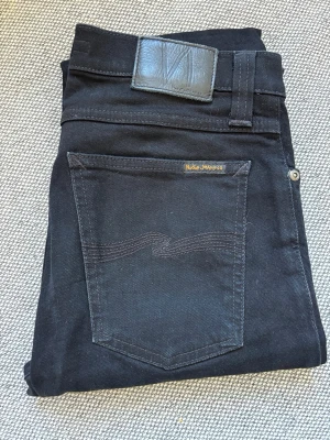Svarta jeans från Nudie Jeans Co - Snygga svarta jeans från Nudie Jeans Co med klassisk femficksdesign och diskret logga på bakfickan. Jeansen har raka ben och är tillverkade i ett mjukt denimtyg. Perfekta för en stilren och avslappnad look.