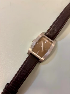 Brun DKNY klocka med läderarmband - Stilren klocka från DKNY med rektangulärt brunt urtavla. Klockan har ett mörkbrunt läderarmband och silverfärgat hölje. Perfekt accessoar för dig som gillar klassisk och tidlös vintage design.