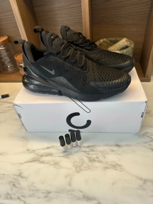Nike Air Max 270 Triple Black sneakers - Snygga Nike Air Max 270 Triple Black sneakers med helsvart mesh- och syntetöverdel, synlig Air Max-dämpning i hälen och klassisk Nike-logga på sidan. Skorna har en sportig siluett och är perfekta för en clean look, perfekt för dig som vill ha snygga skor samtidigt som du gör något sportigt. Box ingår, även 4 mini perfymer från one bold chemist. 