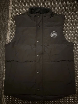 Canada goose väst - Snygg svart dunväst från Canada Goose med hög krage och tryckknappar framtill. Västen har en diskret logotyp på bröstet och är perfekt för lager-på-lager-stil. Klassisk passform och stilren design.