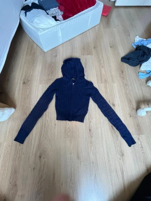 Mörkblå stickad hoodie med dragkedja - Snygg mörkblå stickad hoodie med dragkedja framtill och långa ärmar. Tröjan har ribbade muddar och en åtsittande passform. Perfekt för en chill och stilren look. Materialet är stickat och ger en mysig känsla.