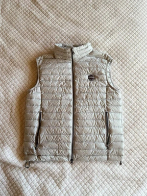 Ljusgrå dunväst från Colmar - Snygg ljusgrå dunväst från Colmar med quiltad design och hög krage. Västen har två dragkedjeförsedda fickor och en rund Colmar-logga på bröstet. Perfekt att slänga över en hoodie eller tröja för extra värme.