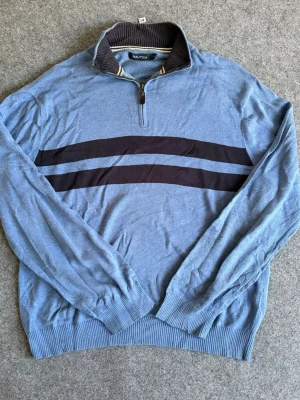 Blå stickad tröja från Nautica XL - Snygg blå stickad tröja från Nautica med två mörkblå ränder över bröstet. Tröjan har halv dragkedja och ribbad krage med vita detaljer. Långa ärmar och ribbade muddar ger en avslappnad och sportig look.