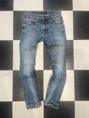 Blå slim fit jeans från Nudie Jeans - Snygga blå slim fit jeans från Nudie Jeans med klassisk femficksdesign och ljusa slitningar på benen. Jeansen har en medelhög midja och tydliga sömmar på bakfickorna. Perfekta för en avslappnad och trendig look. Snygg lagning på höger knä