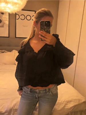 Svart volangblus med v-ringning - Säljer en svart blus med v-ringning och volangdetaljer längs bröstet och axlarna. Blusen har långa ärmar och en luftig, något kortare passform som ger en trendig look. Perfekt att matcha med jeans eller kjol för en cool stil.
