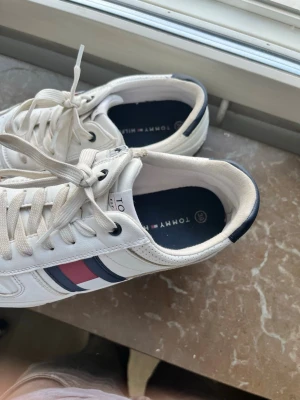 Vita Tommy Hilfiger sneakers - Snygga vita sneakers från Tommy Hilfiger med klassiska blå och röda detaljer på sidan. Skorna har lågt skaft, vita snören och en bekväm sula. Perfekta för en stilren och sportig look. Materialet är mestadels läder och textil.
