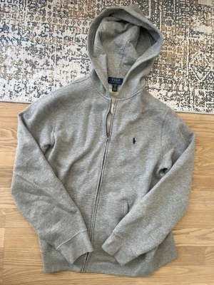 Polo Ralph Lauren Grå hoodie/kofta - Snygg grå hoodie/kofta från Polo Ralph Lauren med dragkedja och klassisk huva. Tröjan har en liten broderad logga på bröstet och är tillverkad i mjukt bomullsmaterial. Perfekt för en avslappnad och stilren look.                                Hoodien är knappt använd