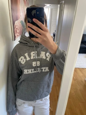 Grå hoodie med tryck 'Paris 95 Atelier' - Snygg grå hoodie med vit text 'PARIS 95 ATELIER' på bröstet. Klassisk modell med huva, dragsko och stor magficka. Perfekt för en avslappnad och trendig stil. Mjuk bomullsblandning som är skön att ha på sig.