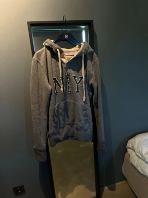 Grå hoodie från G-Rich med NY-tryck - Mysig grå hoodie från G-Rich med stor huva och vita dragsnören. Framtill står det 'NY' i stora bokstäver och 'G-Rich' broderat mellan bokstäverna. Klassisk känguruficka och ribbade muddar. Perfekt för en avslappnad streetwear-look.