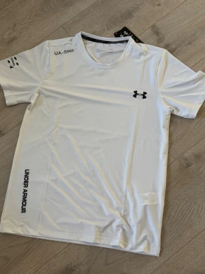 Vit träningströja från Under Armour - Snygg vit träningströja från Under Armour med svart logga på bröstet och tryck på axel och sida. Tillverkad i lätt och snabbtorkande polyester, perfekt för intensiva träningspass. Klassisk rund hals och korta ärmar för maximal rörelsefrihet.