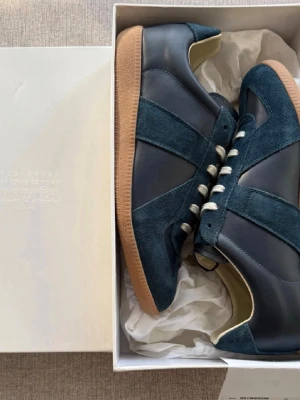 Blå sneakers från Maison Margiela - Snygga sneakers i mörkblått läder och mocka från Maison Margiela. Skorna har vita snören och en klassisk gummisula i brunt. Ikonisk design med diskreta detaljer och låg profil som ger en clean look.