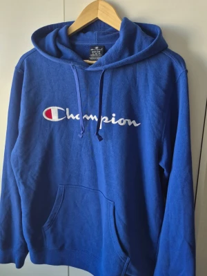 Champion hoodie - Snygg blå hoodie från Champion med stor broderad logga i vitt och rött på bröstet. Klassisk känguruficka framtill och justerbar huva med dragsko. Tillverkad i mjukt bomullsmaterial som är skönt mot huden. Perfekt för en avslappnad och sportig stil.
