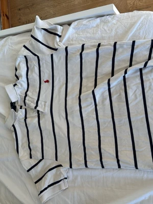  pikè Ralph Lauren - Vit pikétröja från Ralph Lauren med mörkblå horisontella ränder och klassisk krage. Tröjan har korta ärmar, knappar vid halsen och den ikoniska röda logotypen broderad på bröstet. Tillverkad i 100% bomull för en skön och luftig känsla.