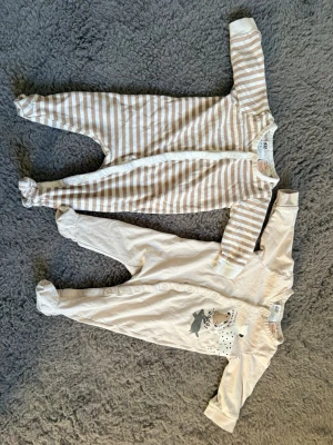 2-pack babypyjamas från H&M, beige/vit - Två mjuka babypyjamas från H&M i bomull. Ena är beige och vitrandig med knäppning framtill, den andra är beige med djurmotiv och knappar längs framsidan. Båda har långa ärmar och täckta fötter, perfekta för mysiga nätter. Minimal användning 