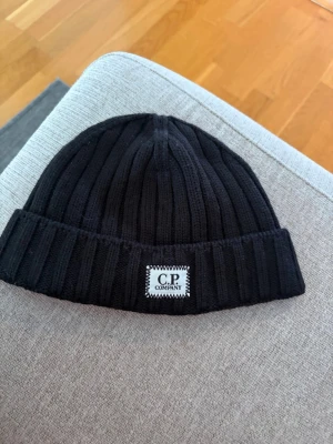 Svart ribbstickad mössa C.P. Company - Svart ribbstickad mössa från C.P. Company med uppvikt kant och vit logopatch framtill. Klassisk och enkel design som passar till många stilar. Materialet är mjukt och värmande, perfekt för kalla dagar.