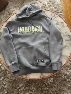 Grå hoodie från Hoodrich med tryck - Snygg grå hoodie från Hoodrich med stor vit och gul logga på bröstet. Klassisk känguruficka framtill och justerbar huva med dragsko. Tillverkad i mjukt bomullsmaterial som är skönt mot huden. Perfekt för en avslappnad streetwear-look.