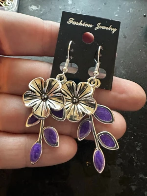 Silvriga blomörhängen med lila blad - Unika örhängen i silverfärgad metall med stora blommor och detaljerade lila blad. Blommorna har en tydlig struktur och örhängena har krokfäste. Perfekta för dig som vill sticka ut med ett färgglatt och kreativt smycke.