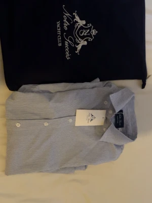 Randig skjorta från Notre Succès - Ny skick - Klassisk blåvit randig skjorta från Notre Succès Yacht Club. Skjortan har knappar framtill, krage och är tillverkad i ett lätt bomullsmaterial. Perfekt för en stilren och avslappnad look.