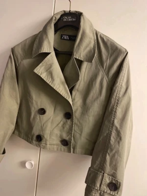 Kort oliv  trenchcoat från Zara - Stilren och croppad trenchcoat från Zara i olivgrön bomullstyg. Jackan har dubbelknäppning med stora knappar, klassisk krage och långa ärmar med knappdetalj vid ärmslut. Perfekt för dig som vill ha en trendig och tidlös look.