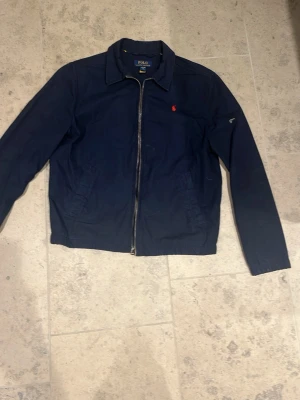 Marinblå jacka från Polo Ralph Lauren - Snygg marinblå jacka från Polo Ralph Lauren med dragkedja framtill och klassisk krage. Jackan har det ikoniska röda Polo-logot broderat på bröstet och två sidofickor. Perfekt för dig som gillar stilren och tidlös design.