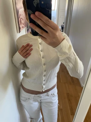 Vit kofta med pärlknappar - Snygg vit kofta med rund halsringning och pärlknappar framtill. Koftan har långa ärmar och breda ribbade muddar vid ärmslut och nederkant. Perfekt att styla med jeans eller kjol för en clean look.