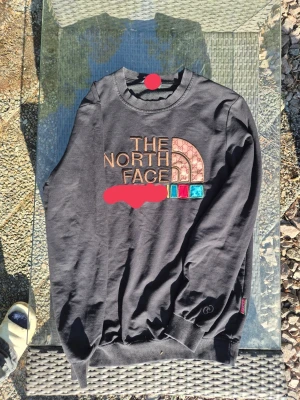 Nort face x gg  - he North Face x gg med stort broderat logotyptryck på bröstet i beige, rött och blått. Tröjan har rund hals, ribbade muddar och en avslappnad passform. Perfekt för dig som gillar streetwear och vill sticka ut.