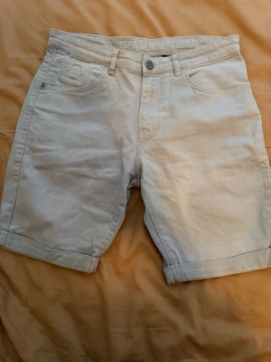 Beige shorts från ISPADENIM - Snygga beige shorts från ISPADENIM med regular fit och klassisk femficksdesign. Shortsen har uppvikta benslut, bälteshällor och knappstängning. Perfekta för varma dagar och enkla att matcha med olika stilar.
