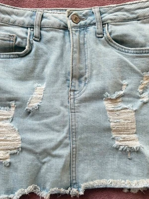 Ljusblå jeanskjol med slitningar - Trendig ljusblå jeanskjol med slitna detaljer och fransig nederkant. Klassisk femficksmodell med knapp och dragkedja framtill. Perfekt för dig som gillar en avslappnad och edgy stil.
