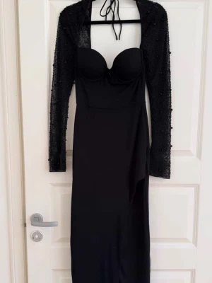 Svart lång klänning med paljettbolero - Snygg svart lång klänning med figurnära passform och bygelkupor. Tillhörande bolero med långa ärmar täckt av svarta paljetter och knytning i nacken. Perfekt för dig som vill sticka ut med en elegant och trendig look.